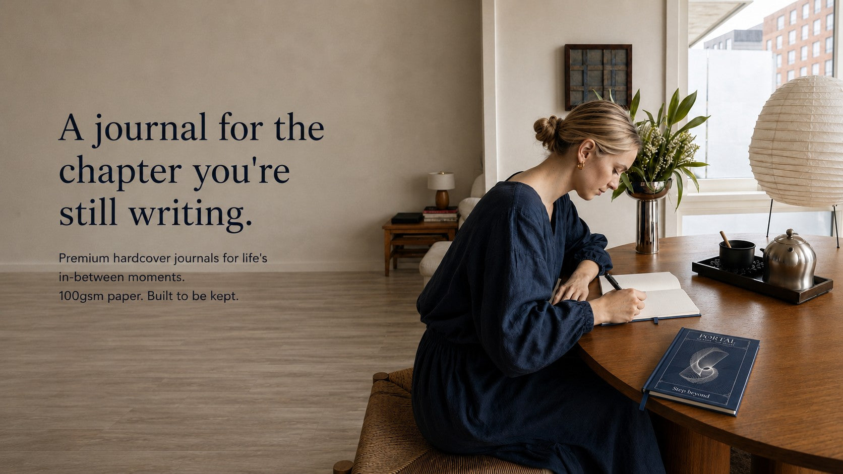 Woman writing in a hardcover journal beside The Ei8ht Portal journal, with soft morning light and calm workspace, for meaningful life transitions.