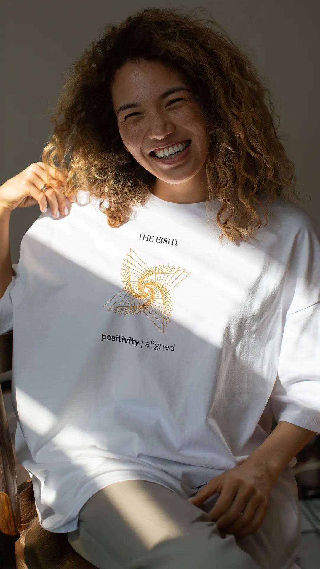 Smiling women wearing oversized white organic cotton t-shirt with orange spiral and "positivity | aligned" message by The Ei8ht, natural light indoor setting.