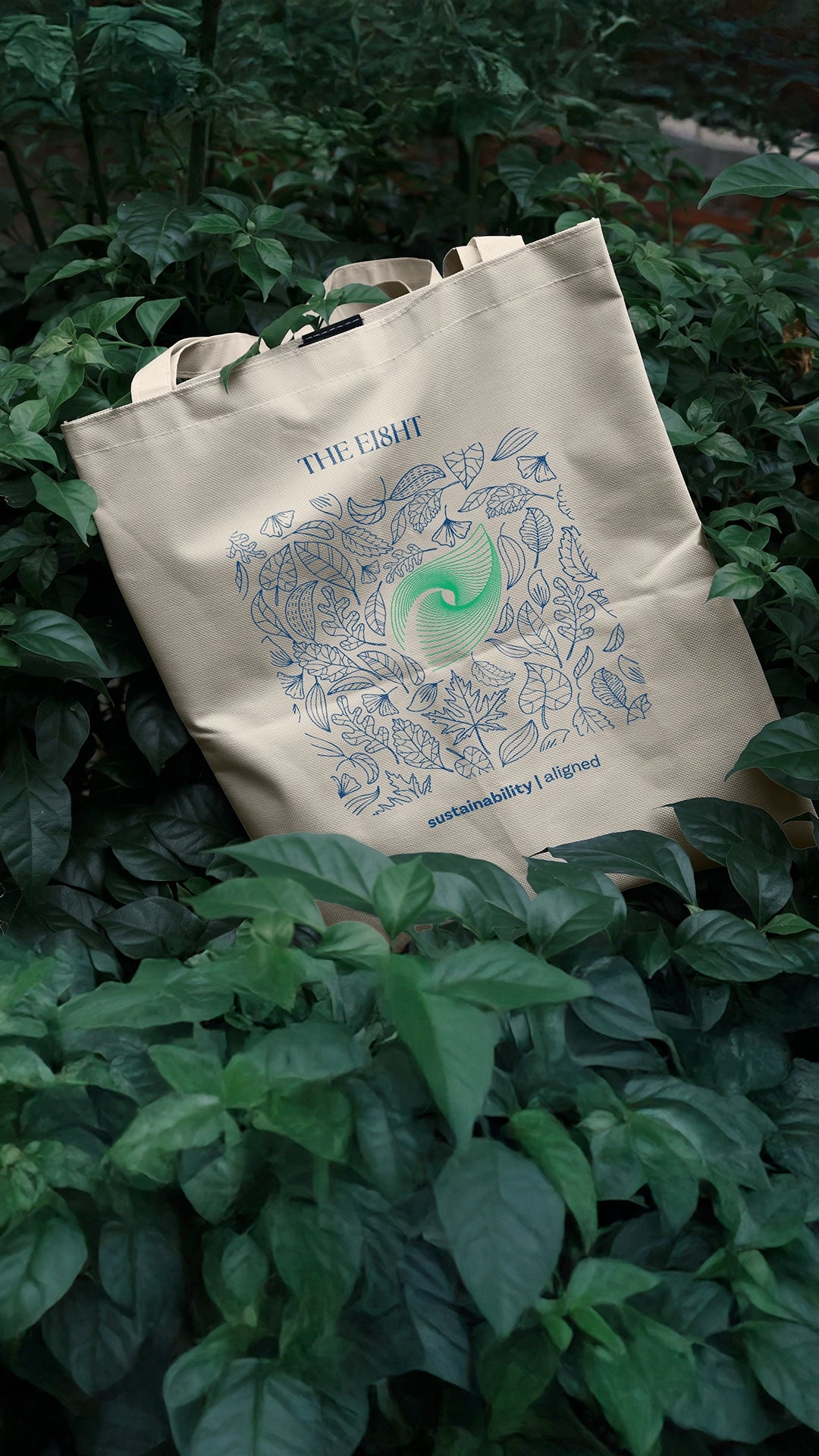 Organic cotton tote bag with green spiral design by The Ei8ht collection, photographed on a lush green leafy background. 